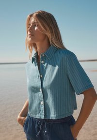 Chemise bleu clair à manches courtes et coupe courte, avec des rayures verticales et une fermeture à boutons à l'avant, associée à un short bleu avec cordons de serrage.