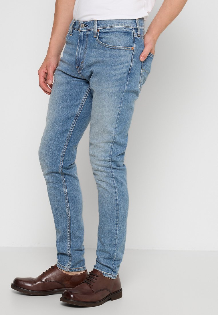 Jeans en denim bleu clair à coupe slim, présentant un design classique à 5 poches et un léger délavage, associés à des chaussures en cuir marron.