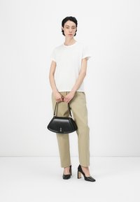 Schwarze Lederhandtasche mit einer strukturierten dreieckigen Form und einem kurzen Griff. Das Modell trägt ein weißes T-Shirt, beige Hosen und schwarze High Heels.