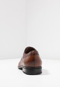 Brune læderherresko med en glat finish, rund tå og teksturerede detaljer på hælen. Sort gummisål. Bagfra.