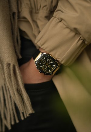 Black watch with a square gold frame, three sub-dials, and a textured black strap, worn on a wrist with a beige garment.