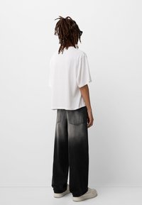 Bílé krátké tričko s krátkým rukávem, volný střih, spárované s černými denimovými kalhotami širokého střihu se zesvětleným vzhledem. Oblečení doplňují béžové tenisky.