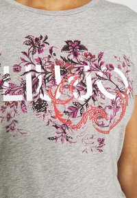 Camiseta de algodón gris con estampado floral en rosa, rojo y negro, con un logo blanco prominente y acentos de cristal para embellecimiento.