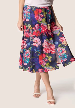 Jupe midi florale en marine, ornée de grandes fleurs roses, rouges et teal. Tissu lisse avec une coupe évasée et une fente sur le côté. Assortie à des talons beiges.