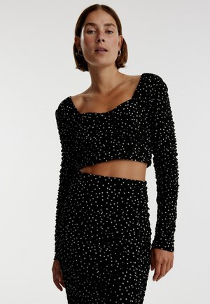 Schwarzes gerafftes zweiteiliges Outfit mit langen Ärmeln, das mit weißen Punkten versehen ist. Das Crop-Top hat einen quadratischen Ausschnitt, der passende Rock hat einen seitlichen Cutout.
