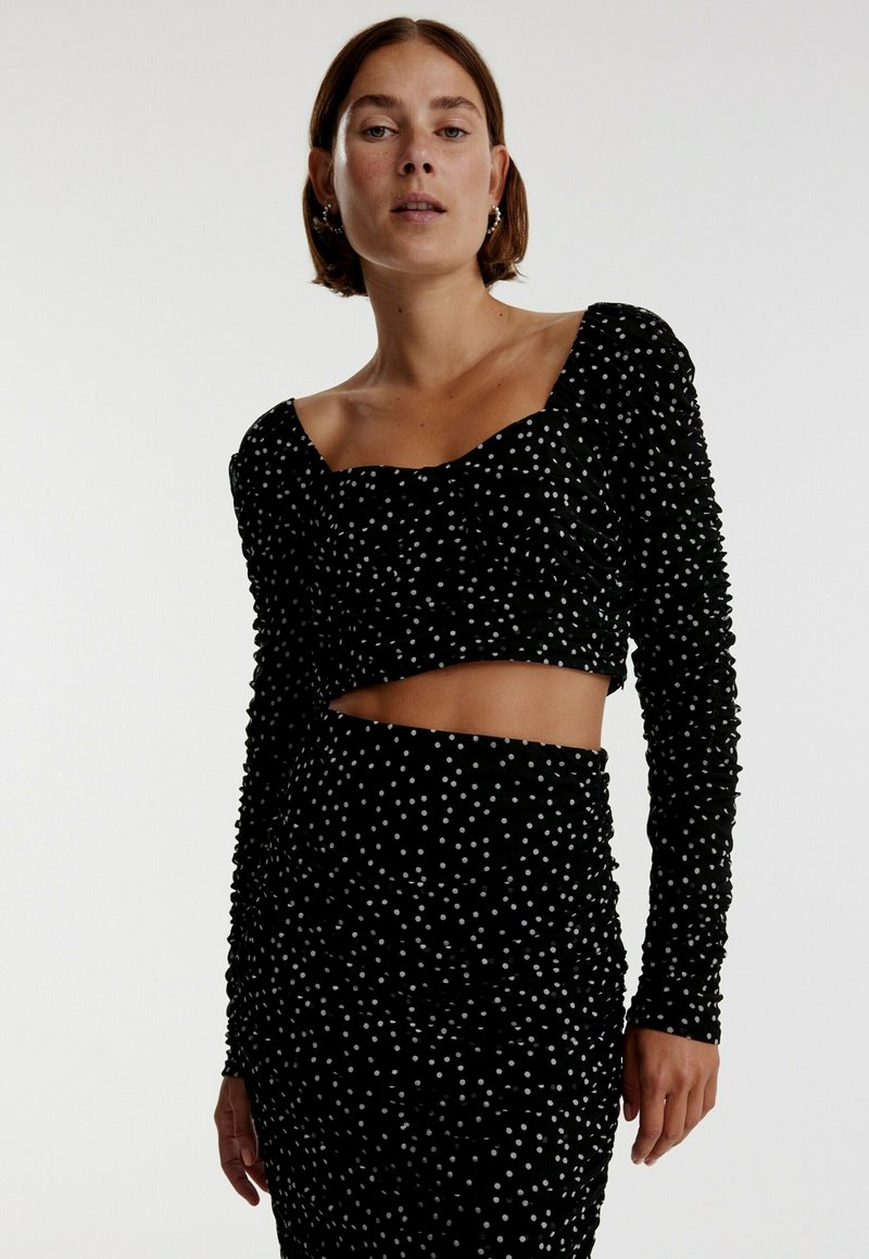 Zwart gerimpeld twee-delig outfit met lange mouwen, met witte stippen. Het crop top heeft een vierkante halslijn en de bijpassende rok heeft een uitsnijding aan de zijkant.