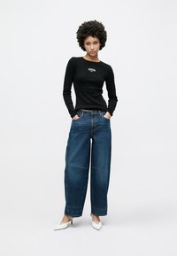 T-shirt preta de manga comprida com logótipo, combinada com jeans azuis de cintura alta e pernas largas, e saltos brancos com bico pontiagudo. Outfit simples e casual.