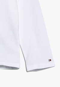 Biała koszula z długim rękawem o gładkiej bawełnianej fakturze. Zawiera małą metkę z logo w kolorach czerwonym, białym i niebieskim blisko dołu. Standardowy krój.