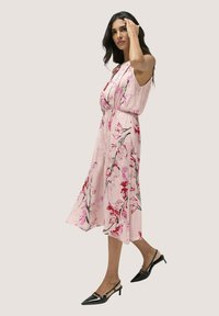 Blumenkleid in lichtrosa Stoff, mit tailliertem Bund und langem, fließendem Rock mit schwarzen und roten Blumenmustern. Schwarze Absatzschuhe.