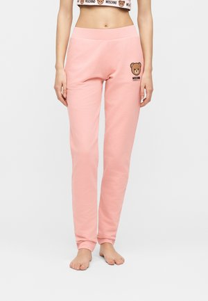 Femme portant un pantalon de jogging rose clair Moschino avec un logo ours en peluche sur la partie supérieure gauche de la cuisse et un crop top assorti.