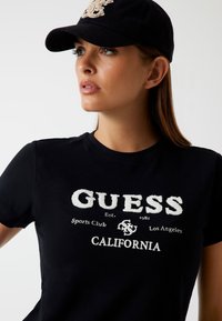 Sort bomulds t-shirt med et hvidt, tekstureret "GUESS" logo og "California" tekst. Sammen med en sort baseball cap.