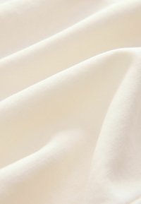 Valkoinen puuvillakangas, jossa on pehmeä ja sileä rakenne, hienovaraisella pintakuvioinnilla, aseteltuna lempeisiin laskoksiin.