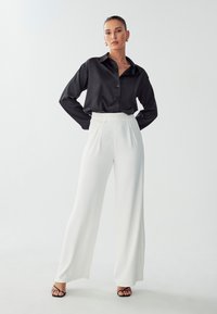 Camicia nera in satin con bottoni e maniche lunghe, abbinata a pantaloni bianchi a gamba larga con vita alta e pieghe frontali. Sandali neri con cinturini.