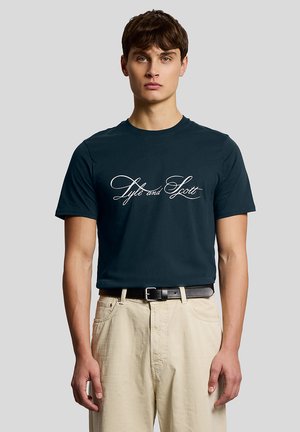 Junger Mann trägt ein marineblaues T-Shirt mit dem Schriftzug "Lyle and Scott" in Kursivschrift, beige Hose und schwarzen Gürtel, steht vor einem schlichten Hintergrund.