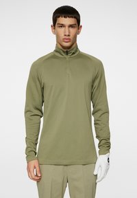 Olivgrön zip-up långärmad topp i ett lätt, strukturerat tyg med ett subtilt mönster; matchad med ljusa byxor och en vit handske.