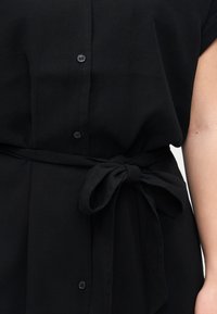 Μαύρο πουκάμισο με κουμπιά και υφασμάτινη ζώνη δεμένη σε φιόγκο στη μέση, που αποκαλύπτει μερικώς γυμνό βραχίονα στη δεξιά πλευρά.