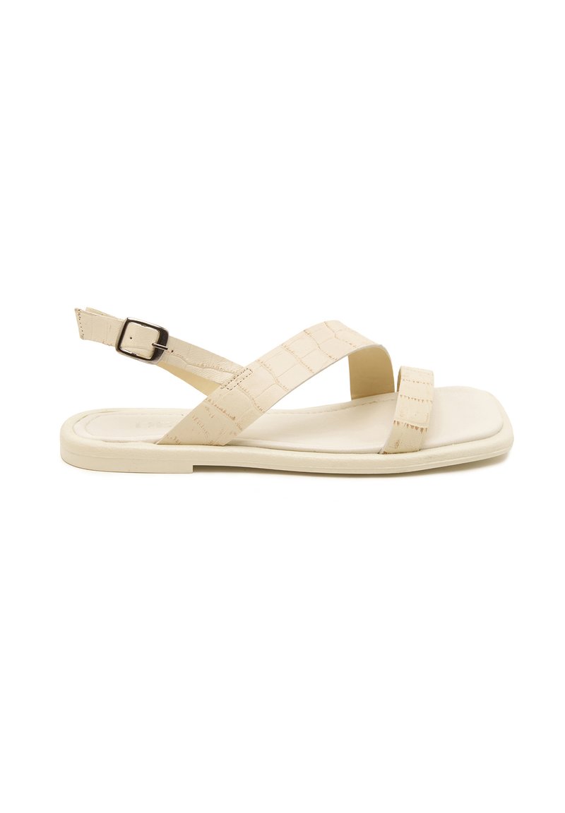 Beige sandal with cross straps and back buckle; features a smooth, flat sole and a subtle croc pattern for added texture.