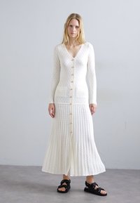 Vrouw in een lange witte gebreide knoopjurk met zakken, staande op een grijze vloer tegen een effen lichtgrijze muur, met zwarte sandalen.
