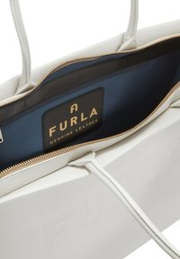 Sac fourre-tout en cuir blanc avec une fermeture éclair dorée. L'intérieur présente une doublure bleue et une étiquette noire imprimée "FURLA CUIR AUTHENTIQUE."