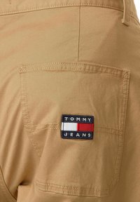 Bolsillo de pantalones beige con parche rectangular de logo de Tommy Jeans en negro, blanco, rojo y azul marino, cosido en la tela.