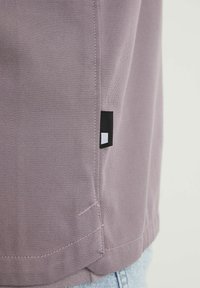 Gros plan de la couture latérale d'une chemise violette avec une petite étiquette en tissu noir et blanc près de l'ourlet au-dessus d'un jean bleu clair.