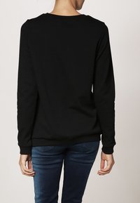 Černá mikina s dlouhými rukávy, kulatým výstřihem, měkkou texturou a žebrovanými manžetami a lemem, v kombinaci s modrými džínovými kalhotami.