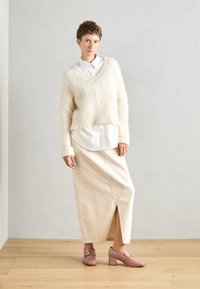 Cremfarbener, flauschiger Pullover mit V-Ausschnitt, der über einem weißen Hemd getragen wird. Beigefarbener Rock mit Schlitz vorne und rosa Blockabsatzschuhen.