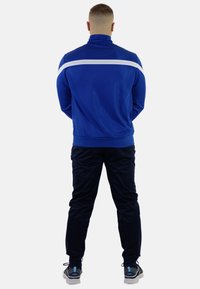 Blå atletisk jakke med høj krave og hvid stribe over skuldrene, parret med sorte bukser og sneakers, set bagfra.