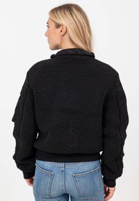 Giubbotto bomber nero, texture a pelo, con finitura pelosa, polsini e orlo a costine, e colletto alto, visto da dietro.