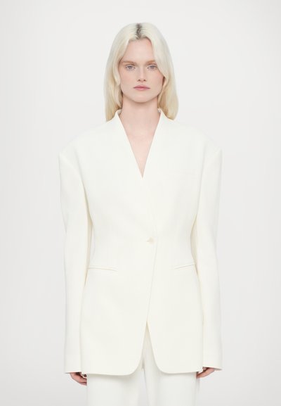 Blazer blanc avec un design de revers net, coupe oversize, fermeture à un bouton et texture élégante. Associé à un pantalon assorti.