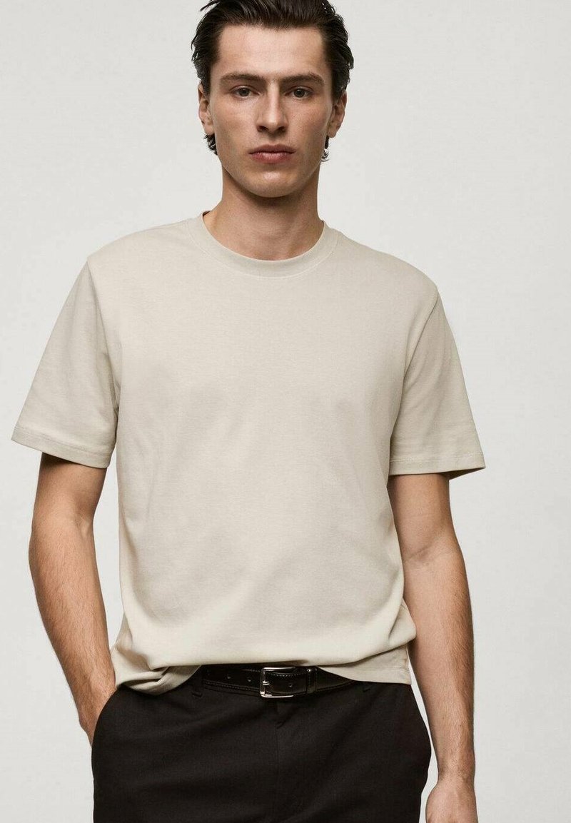 Maglietta in cotone beige con scollatura rotonda e maniche corte, caratterizzata da una texture liscia. Indossata con pantaloni neri e una cintura.
