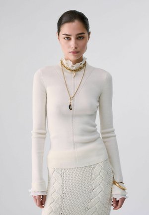 Femme aux cheveux foncés portant un pull côtelé crème et une jupe en maille torsadée assortie, avec des colliers et bracelets dorés superposés sur un fond uni.