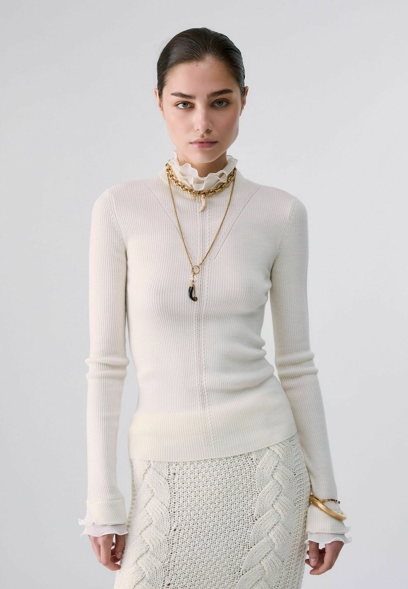 Femme aux cheveux foncés portant un pull côtelé crème et une jupe en maille torsadée assortie, avec des colliers et bracelets dorés superposés sur un fond uni.