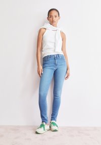 Femme se tenant contre un mur blanc, portant un top sans manches blanc, un sweat-shirt blanc noué autour des épaules, un jean skinny bleu et des baskets blanches avec des lacets verts.