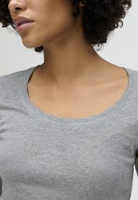 Woman with curly hair wearing a light grey round-neck sweater, shown from the chin to the upper chest against a plain background.
