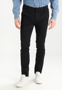 Černé slim-fit kalhoty vyrobené z elastické tkaniny, s klasickým pětipocket designem, v kombinaci s bílými teniskami a světlou džínovou košilí.