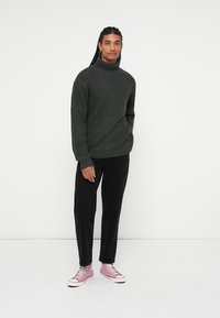 Mørkegrøn strikket sweater med høj krave, sorte bukser og lyserøde high-top sneakers. Outfittet har en afslappet pasform og en struktureret materiale.