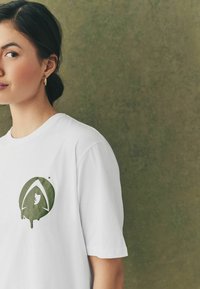 Hvid bomulds t-shirt med et grønt grafisk logo med dråbe-design på venstre brystområde. Korte ærmer og rund hals.