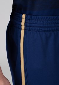 Shorts de sport navy avec taille élastique, dotés d'une bande dorée sur le côté. Le matériau est lisse et léger avec une finition texturée.