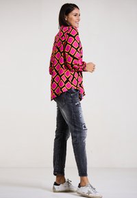 Mehrfarbige Bluse mit einem Diamantmuster in Pink, Gelb und Schwarz, kombiniert mit verwaschenen grauen Jeans und weißen Sneakers.