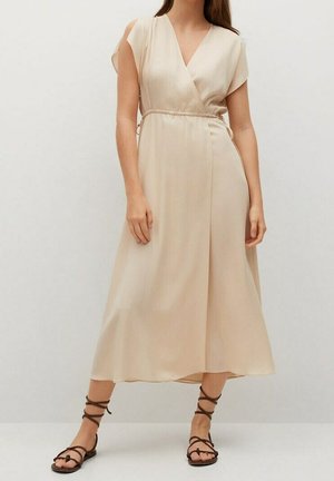 Kobieta ubrana w beżową, bezrękawnikową sukienkę midi z kopertowym wiązaniem w talii oraz ciemnobrązowe, sznurowane płaskie sandały, stojąca na jednolitym tle.