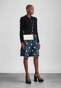 Femme portant un haut noir à manches longues, une jupe en jean brodée de fleurs, des sandales à plateforme noires et un sac bandoulière blanc, debout devant un fond gris.