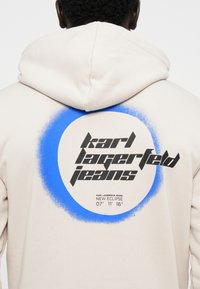Felpa beige con un grande cerchio blu spruzzato e testo nero in grassetto che recita "karl lagerfeld jeans" e "NEW ECLIPSE" sul retro.