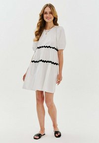 Biele krátke šaty s krátkym rukávom a okrúhlym výstrihom, s čiernym vlnitým lemovaním. Voľný strih s rozšírenou siluetou. Nosené s čiernymi šľapkami.