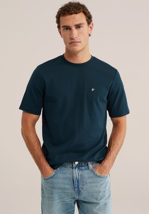 Man met krullend haar, die een donkerblauw T-shirt met korte mouwen en lichtblauwe spijkerbroek draagt met handen in de zakken, neutrale achtergrond.