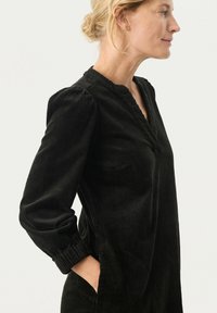 Robe en velours côtelé noir avec un col mandarin, des manches longues, des poignets froncés et des poches latérales ; le tissu texturé crée un effet de côtes verticales.