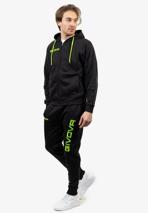 Uomo che indossa un felpa nera con zip e pantaloni abbinati con il brillante marchio verde "GIVOVA", in piedi con le mani nelle tasche, sneakers bianche.