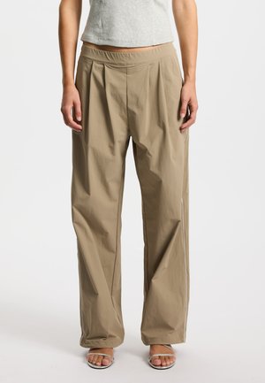 Beige, weite Hosen mit glatter Textur, elastischem Bund und seitlicher Paspelierung. Kombiniert mit grauem Oberteil und silbernen Sandalen.