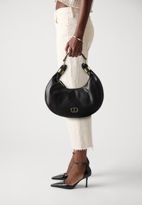 Black leather handbag with a curved shape, chunky gold hardware, and a smooth texture. Worn with light-colored pants and black pointed heels.