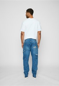 Blaue Denim-Cargohosen mit mehreren Taschen, verziert mit bestickten Details und einem lockeren Schnitt. Kombiniert mit einem schlicht weißen T-Shirt.